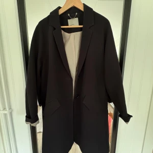 Svart kappa från Vero Moda - Stilren svart kappa från Vero Moda med klassisk krage och två knappar framtill. Jackan har långa ärmar, två fickor och är helfodrad med ljus insida. Perfekt för dig som gillar en enkel och tidlös look.