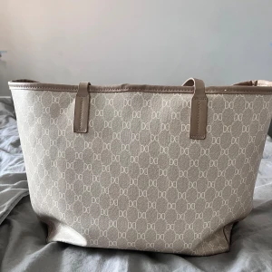 Beige shoppingväska från Don Dona - Säljer en beige shoppingväska från Don Dona med klassiskt monogrammönster och ljusbruna detaljer. Väskan har två handtag och rymligt huvudfack, perfekt för att bära med sig allt man behöver. Snygg och stilren design!