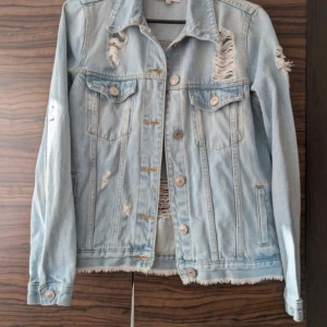 Ljusblå jeansjacka från River Island - Säljer en ljusblå jeansjacka från River Island med slitna detaljer och knappar framtill. Jackan har två bröstfickor med lock och långa ärmar. Perfekt för dig som gillar en trendig och avslappnad stil.