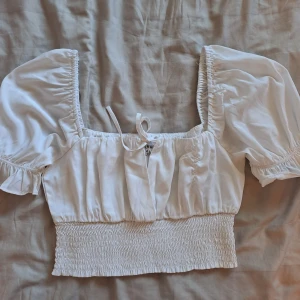 Vit croppad blus med puffärmar och knyt - Säljer en söt vit croppad blus från NA-KD med puffiga korta ärmar, smockad midja och knyt framtill. Blusen har en fyrkantig urringning och volangdetaljer vid ärmslut. Perfekt för vår och sommar! Den är köpt secondhand och har flera slitningar (bild 3). Kan tänka mig att sänka priset ännu mer, vill mest få bort den ur min garderob❤️