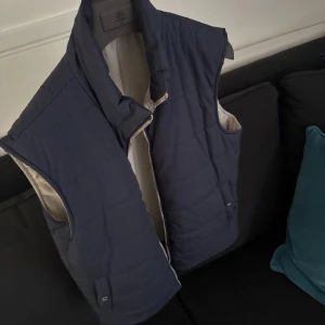 Mörkblå quiltad dunväst från Eleventy - Stilren mörkblå quiltad dunväst från Eleventy med dragkedja och två fickor framtill. Insidan är ljusbeige och västen har en hög krage. Perfekt för lager-på-lager under kyliga dagar. Ny pris runt 12k.