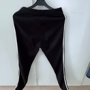 Svarta leggings med vita ränder - Svarta leggings med vita ränder längs sidorna. Byxorna har en tight passform och elastisk midja, perfekta för en sportig och avslappnad stil.