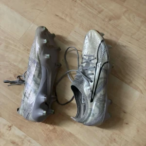 Silvriga fotbollsskor från Puma - Säljer ett par silvriga fotbollsskor från Puma med snörning och rund tå. Skorna har en lätt och smidig design med platt sula och dobbar för spel på gräs. Perfekta för dig som vill ha snabba fötter på planen!
