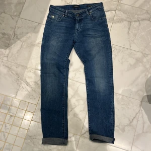 Blå jeansbyxor från Scotch & Soda - Säljer ett par klassiska blå jeans från Scotch & Soda, modell Strummer. Jeansen har normal passform, raka ben och snygga detaljer på bakfickan. Perfekta till vardags och har en stilren look med uppvikta benslut. Storlek 152 
