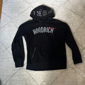 Svart hoodie från Hoodrich med broderade detaljer - Säljer en svart hoodie från Hoodrich med stora broderade loggor i vitt och rött på bröstet och huvan. Hoodien har långa ärmar och en klassisk passform. Perfekt för dig som gillar streetwear och snygga detaljer.