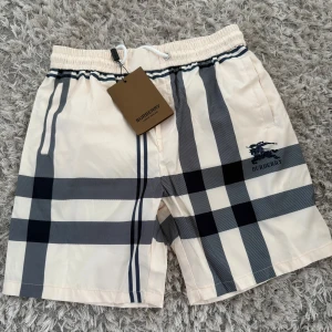 Burberry rutig shorts med snörning - Snygga Burberry shorts i klassiskt beige med stora svarta och grå rutor. Shortsen har elastisk midja med vit snörning och sidofickor. Tillverkade i Italien och märkta med Burberry-logga på benet. Perfekta för sommaren.