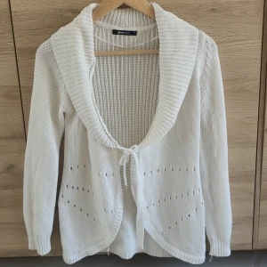 Vit stickad kofta från Gina Tricot - Säljer en vit stickad kofta från Gina Tricot med lång ärm, bred krage och knytband framtill. Koftan har ett diskret hålmönster och ribbade muddar vid ärmslut och nederkant. Perfekt för lager-på-lager-stil!