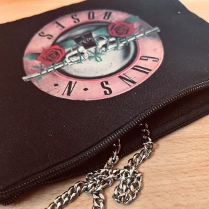 Svart necessär i canvas med Guns n' Roses-motiv! - Svart necessär i canvas med Guns n' Roses-motiv framtill och dragkedja. Mått: 20 x 22 cm. Oanvänd.
