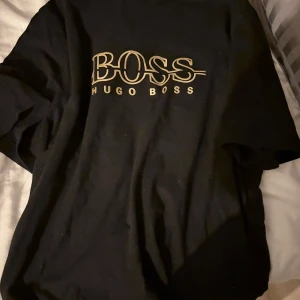 Svart t-shirt från Hugo Boss - Säljer en svart t-shirt från Hugo Boss med stor guldfärgad logga på bröstet. Klassisk passform och rund halsringning. Perfekt för dig som gillar stilrena märkesplagg.