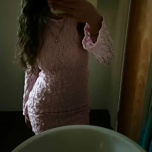 Ljusrosa figurnära klänning med struktur - Säljer en ljusrosa klänning med figurnära passform och strukturerat tyg. Klänningen har långa ärmar med volangdetalj vid ärmslut och hög hals och öppen rygg. Perfekt för dig som vill sticka ut med en feminin och trendig look.