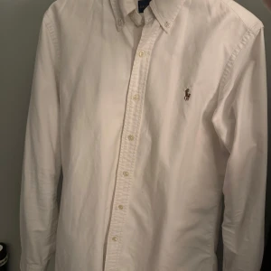 Vit skjorta från Ralph Lauren - Klassisk vit skjorta från Ralph Lauren med broderad logga på bröstet. Skjortan har lång ärm, knappar framtill och en stilren krage. Perfekt för en clean och tidlös look.