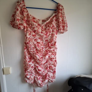 Blommig kortklänning med puffärm - Söt kortklänning med röd-rosa blommönster på vit botten. Klänningen har puffiga korta ärmar, snörning framtill och lätt rynkade detaljer. Perfekt för sommarens alla tillfällen.
