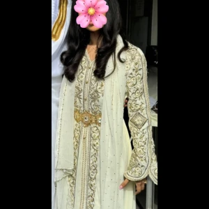 Vit / guldig kaftan  - Elegant kaftan med detaljerade guldiga broderier och pärlor. Klänningen har långärmade ärmar och ett brett dekorerat bälte i midjan. Materialet ser ut att vara syntet eller polyester med glittriga inslag och vackra mönster längs hela klänningen. Använd endast i 1 timme på min Nikkah. 