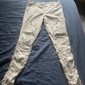 Beige cargopants från Sub Culture - Snygga beige cargopants från Sub Culture med flera praktiska fickor och dragkedjor. Byxorna har en modern design med rynkade detaljer längs benen och en bekväm passform. Perfekta för en avslappnad stil.