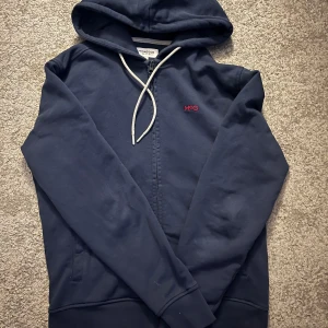 Mörkblå hoodie från McGregor - Säljer en snygg mörkblå hoodie från McGregor med dragkedja och broderad logga i rött på bröstet. Den har en justerbar huva med vita snören och långa ärmar. Perfekt för en avslappnad stil.