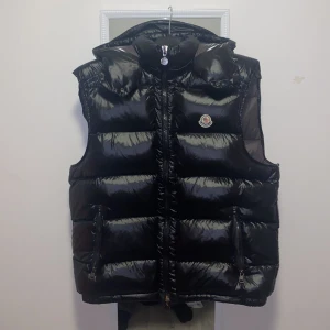 Svart dunväst från Moncler - Snygg svart dunväst från Moncler med glansig yta och dragkedja framtill. Västen har en broderad Moncler-logga på bröstet och praktiska fickor med dragkedja. Perfekt för kyligare dagar.