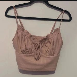 Rosa ribbad linnetopp - Säljer en söt rosa ribbad linnetopp med smala axelband och knytning framtill. Perfekt för varma sommardagar eller som ett lager under en jacka. Toppen har en croppad passform som ger en trendig look.