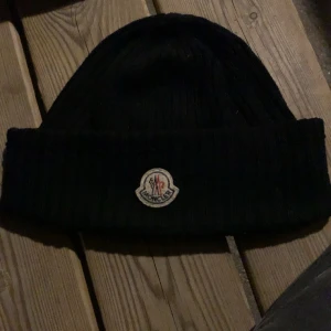 Svart ribbad mössa från Moncler - Snygg svart mössa från Moncler med ribbad design och uppvikt kant. Perfekt för att hålla värmen under kyliga dagar. Den har en broderad Moncler-logga framtill.