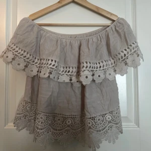 Beige offshoulder topp med spetsdetaljer - Charmig beige offshoulder topp med vackra spetsdetaljer och volanger. Perfekt för en somrig look med sin luftiga design och romantiska stil. Pris kan diskuteras 