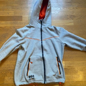 Grå hoodie från Helly Hansen - Säljer en grå hoodie från Helly Hansen med orange detaljer och dragkedja. Jackan har en justerbar huva och två fickor med dragkedjor. Perfekt för en sportig look!