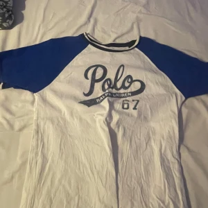Vit och blå t-shirt från Ralph Lauren - Snygg vit t-shirt med blå ärmar från Ralph Lauren. Den har ett stort 'Polo' tryck på framsidan och är perfekt för en avslappnad stil. Klassisk design med rund halsringning.