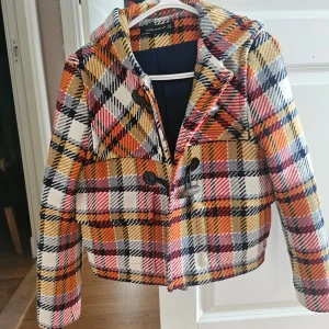Rutig jacka från Zara - Säljer en färgglad rutig jacka från Zara med huva och stora knappar. Jackan har en mix av orange, rött, svart och vitt i mönstret. Perfekt till våren och hösten. Mycket fint skick. 