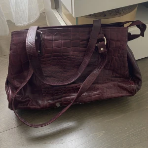 Mörklila axelväska i syntet - Snygg mörklila axelväska med krokodilmönster och dragkedja. Väskan har dubbla handtag och en justerbar axelrem för extra bekvämlighet. Perfekt för att bära med sig allt man behöver under dagen. Lite slitet se bilder❤️ Vintage väska