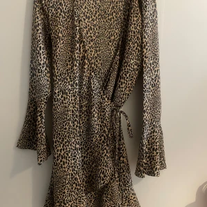 Leopardmönstrad omlottklänning - Trendig leopardmönstrad omlottklänning med knytning i midjan. Klänningen har långa ärmar och en stilren design som passar perfekt för en modemedveten look.