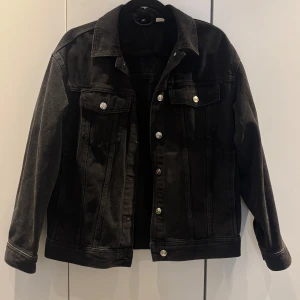 Svart jeansjacka med knappar - Snygg svart jeansjacka med silverfärgade knappar. Klassisk design med två bröstfickor och krage.