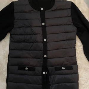  cardigan jacka liknar moncler - Snygg svart quiltad cardigan jacka med långa ärmar och knappar framtill. Jackan har en stilren design med ribbade detaljer vid ärmslut och nederkant. Perfekt för kyligare dagar. Passar s/m