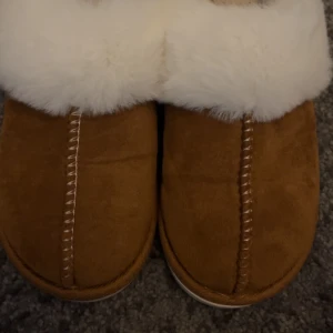 Bruna tofflor med vit päls från lager 157 - Mysiga bruna tofflor med vit päls runt kanten. Perfekta för att hålla fötterna varma under kalla dagar. Tofflorna har en mjuk insida och en stabil sula.