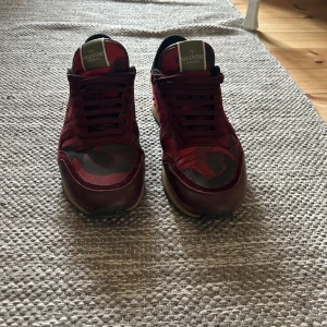 Valentino Rockrunner sneakers i röd camouflage - Snygga Valentino Rockrunner sneakers i en cool röd och svart camouflage-design. Skorna har snörning och en unik detalj med nitar på hälen. Perfekta för att ge din outfit en edgy touch.