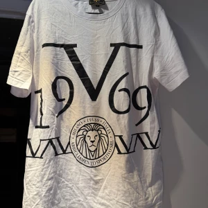 Vit t-shirt från 19V69 Italia - Säljer en stilren vit t-shirt från 19V69 Italia med ett stort svart tryck av lejon och texten 'Milano 1969'. Perfekt för en casual look med en touch av italiensk design. 🦁