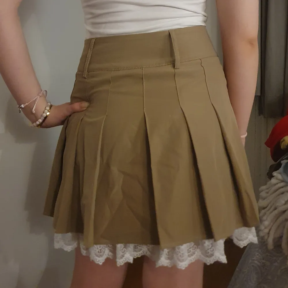 Snygg beige kortkjol med veckad design och vit spetskant som sticker fram nedtill. Kjolen har knappar framtill och en stilren look som passar perfekt för olika tillfällen.. Hameet.