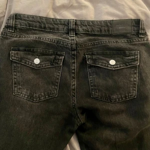 Mörkgrå lågmidjade butcut jeans - Snygga svarta jeansbyxor med klassisk femficksdesign och knappstängning. Byxorna är butcut och har en lågmidjad passform med knappfickor. De har också dekorativa sömmar och en bekväm midja. 