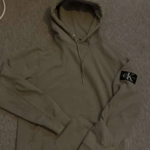  hoodie från Calvin Klein - Säljer en stilren hoodie från Calvin Klein med en klassisk logga på ärmen. Tröjan har en justerbar huva med dragsko och långa ärmar. Perfekt för en avslappnad look.