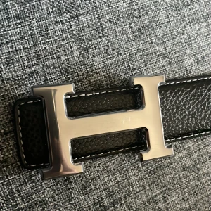 Svart skärp med H-spänne äkta  - Snyggt svart skärp i skinn med ett stort H-format spänne i silver. Skärpet har en stilren design med vita sömmar längs kanterna och flera hål för justering. Perfekt för att ge en elegant touch till din outfit.