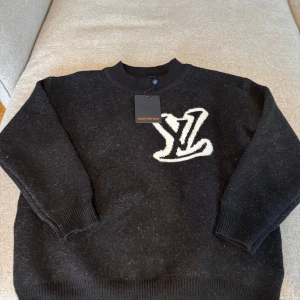 Svart stickad tröja från Louis Vuitton - Säljer en svart stickad tröja från Louis Vuitton med ett stort vitt LV-märke på framsidan. Tröjan har långa ärmar och en rund halsringning. Perfekt för en stilren och lyxig look.
