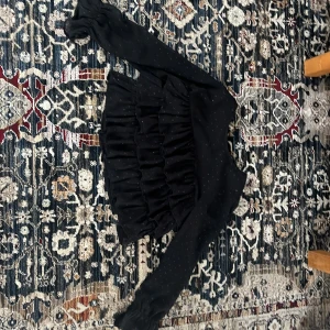 Svart volangblus med prickar - Snygg svart blus med volanger och små prickar. Blusen har långa ärmar och en elegant design som passar perfekt för en kväll ute. Den är tillverkad i ett lätt och bekvämt material.