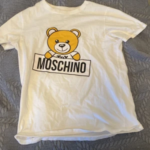 Vit t-shirt med nalletryck från Moschino - Säljer en vit t-shirt från Moschino med ett gulligt nalletryck och texten 'Toy Moschino' på framsidan. Perfekt för en avslappnad stil med en touch av lyx. T-shirten har korta ärmar och är gjord i mjukt bomullsmaterial.