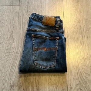 Blå jeans från Nudie Jeans - Snygga blå jeans från Nudie Jeans med klassisk femficksdesign och orange sömmar. Jeansen har en straight passform och är tillverkade i slitstarkt denim. Perfekta för en avslappnad stil.