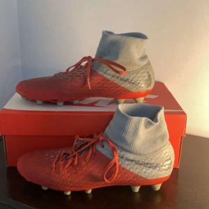 Röda fotbollsskor från Nike - Snygga röda fotbollsskor från Nike med grå detaljer och snörning. Skorna har en hög ankelkrage för extra stöd och en mönstrad yta för bättre grepp på planen. Perfekta för fotbollsspelare som vill ha stil och funktionalitet.