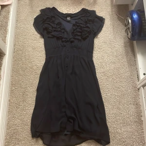 Mörk blå klänning från H&M - Säljer en elegant svart klänning från H&M med volangdetaljer vid halsringningen. Klänningen har korta ärmar och knappar framtill. Perfekt för en stilren look.