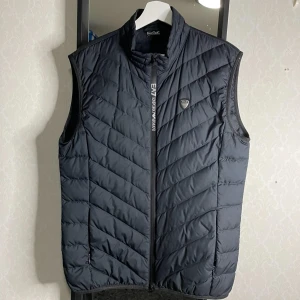Dunväst från Emporio Armani - Snygg svart dunväst från Emporio Armani med dragkedja och ett stilrent märke på bröstet. Perfekt för lager-på-lager under kyliga dagar. Västen har en quiltad design som ger en modern look. Säljes då det inte är min stil längre. Priset kan diskuteras!