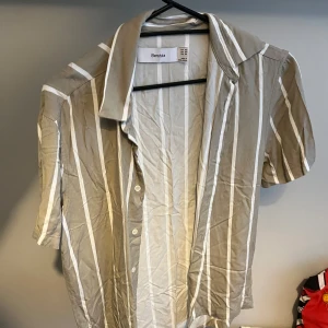 Beige och vit randig skjorta från Bershka - Säljer en stilren beige och vit randig skjorta från Bershka. Skjortan har korta ärmar och knappar framtill. Perfekt för en avslappnad look.