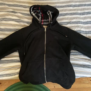 Svart Burberry hoodie med dragkedja - Snygg svart hoodie med dragkedja och rutigt foder i huvan. Perfekt för en avslappnad stil med praktiska fickor och justerbar huva.
