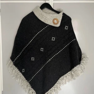 Svart och grå poncho med fransar - Säljer en stilren svart och grå poncho i ull blandning med dekorativa fransar och ett snyggt mönster. Ponchon har en stor krage med en träknapp som detalj. 