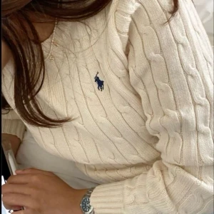 Beige stickad tröja från Ralph Lauren - Säljer en stilren beige stickad tröja från Ralph Lauren med kabelstickat mönster och den klassiska loggan på bröstet. Tröjan har långa ärmar och rund halsringning. Perfekt för en avslappnad och elegant look.