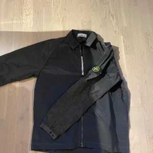 Svart windbreaker från Stone Island - Svart windbreaker från Stone Island med klassisk krage och hel dragkedja framtill. Jackan har lång ärm med den ikoniska loggan på vänster arm och är tillverkad i ett lätt, syntetmaterial. Perfekt för lager-på-lager 