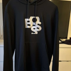 Svart hoodie från BOSS med brodyr - Svart hoodie från BOSS med stor broderad BOSS-logga i vit på bröstet. Klassisk huva med snörning och känguruficka framtill. Tillverkad i mjuk bomullsmix, perfekt för en chill och stilren look. Aldrig använd då den varit för stor, bara hängt och tagit plats i garderoben